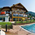 Das mehrstöckige Hotel mit Balkonen überblickt einen eingezäunten Swimmingpool im Freien, umgeben von Gras, Sonnenschirmen und Liegestühlen in der Nähe eines bewaldeten Hügels.