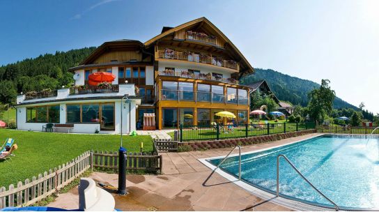 Das mehrstöckige Hotel mit Balkonen überblickt einen eingezäunten Swimmingpool im Freien, umgeben von Gras, Sonnenschirmen und Liegestühlen in der Nähe eines bewaldeten Hügels.