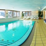 Hallenbad mit gefliestem Boden, großen Fenstern, die eine verschneite Landschaft zeigen, weißen Stühlen und einem Whirlpool auf der rechten Seite.