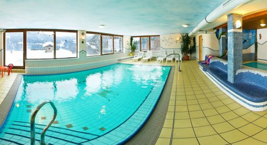 Hallenbad mit gefliestem Boden, großen Fenstern, die eine verschneite Landschaft zeigen, weißen Stühlen und einem Whirlpool auf der rechten Seite.