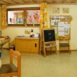 Ein hell erleuchtetes Klassenzimmer mit farbenfroher Dekoration, Kinderkunstwerken an den Wänden, Tischen, Stühlen, Spielzeug und einer Staffelei mit einer Kreidezeichnung.