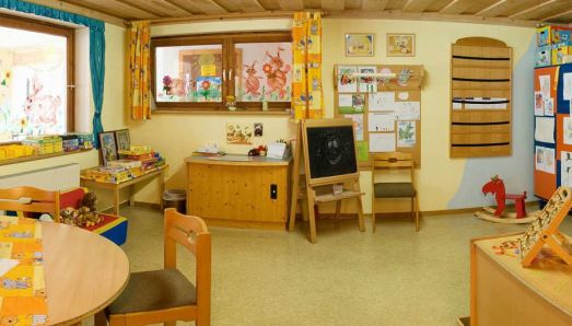 Ein hell erleuchtetes Klassenzimmer mit farbenfroher Dekoration, Kinderkunstwerken an den Wänden, Tischen, Stühlen, Spielzeug und einer Staffelei mit einer Kreidezeichnung.