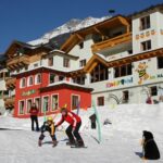 Zwei Erwachsene und zwei Kinder fahren auf einer verschneiten Piste vor einem farbenfrohen Alpenhotel, umgeben von Schnee, mit Bergen im Hintergrund.