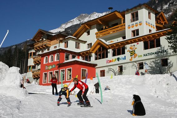 Zwei Erwachsene und zwei Kinder fahren auf einer verschneiten Piste vor einem farbenfrohen Alpenhotel, umgeben von Schnee, mit Bergen im Hintergrund.