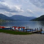 Eine ruhige Szene am Seeufer mit angedockten Paddelbooten, sanft plätscherndem Wasser und Bergen im Hintergrund unter einem bewölkten Himmel.