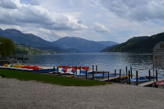 Eine ruhige Szene am Seeufer mit angedockten Paddelbooten, sanft plätscherndem Wasser und Bergen im Hintergrund unter einem bewölkten Himmel.