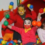 Vier Kinder mit bemalten Gesichtern spielen in einer Ballgrube, lächeln und halten bunte Plastikbälle in der Hand, während im Hintergrund eine rote Tunnelrutsche zu sehen ist.