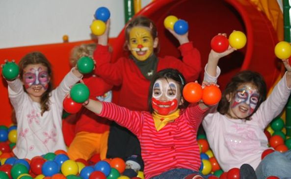 Vier Kinder mit bemalten Gesichtern spielen in einer Ballgrube, lächeln und halten bunte Plastikbälle in der Hand, während im Hintergrund eine rote Tunnelrutsche zu sehen ist.