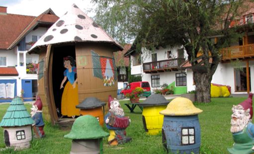 Eine kleine, mit Schneewittchen und einem Zwerg bemalte Hütte steht auf einer grasbewachsenen Wiese, umgeben von bunten Zwergenstatuen und nahe gelegenen Gebäuden.