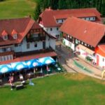 Luftaufnahme einer ländlichen Anlage mit roten Gebäuden, Sitzgelegenheiten im Freien unter Sonnenschirmen und einer von Bäumen umgebenen Rasenfläche.