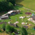 Luftaufnahme eines Spielplatzes im Freien mit verschiedenen Spielgeräten, Trampolinen, Schaukeln und kleinen Holzgebäuden auf einer Wiese in der Nähe eines Waldgebiets.