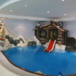 Kinderhallenbad mit einem verschneiten Bergthema, mit einem Bären auf Skiern, einer Holzhütte, einer roten Rutsche und dekorativem Schnee und Tieren.