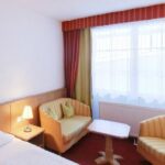 Ein kleines Hotelzimmer mit einem Bett, einem Nachttisch mit einer Lampe, zwei Sesseln, einem Holztisch und großen Fenstern mit durchsichtigen Vorhängen und gestreiften Gardinen.