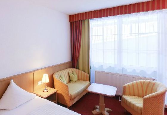 Ein kleines Hotelzimmer mit einem Bett, einem Nachttisch mit einer Lampe, zwei Sesseln, einem Holztisch und großen Fenstern mit durchsichtigen Vorhängen und gestreiften Gardinen.