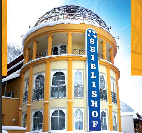 Ein rundes, gelbes Gebäude mit gewölbten Fenstern, einem Kuppeldach und einem vertikalen blauen Schild mit der Aufschrift "Seiblishof", mit Schnee auf dem Dach.