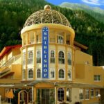 Gelbes Hotelgebäude mit einem Kuppeldach und einem großen blauen vertikalen Schild mit der Aufschrift "SEIBLISHOF" vor einem Hintergrund aus grünen Bäumen und Bergen unter blauem Himmel.