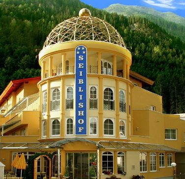 Gelbes Hotelgebäude mit einem Kuppeldach und einem großen blauen vertikalen Schild mit der Aufschrift "SEIBLISHOF" vor einem Hintergrund aus grünen Bäumen und Bergen unter blauem Himmel.