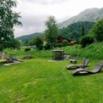 Ein grasbewachsener Hof mit Liegestühlen, einer Bank, einem Pavillon und Bäumen vor der Kulisse von Hügeln und Bergen unter einem bewölkten Himmel.