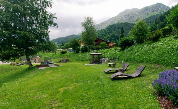 Ein grasbewachsener Hof mit Liegestühlen, einer Bank, einem Pavillon und Bäumen vor der Kulisse von Hügeln und Bergen unter einem bewölkten Himmel.