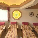 Eine Reihe von hölzernen Lounge-Sesseln mit karierten Kissen und roten Kopfstützen steht in einem gemütlichen Raum mit dekorativer Wandkunst, Vorhängen und einem Buntglasfenster.