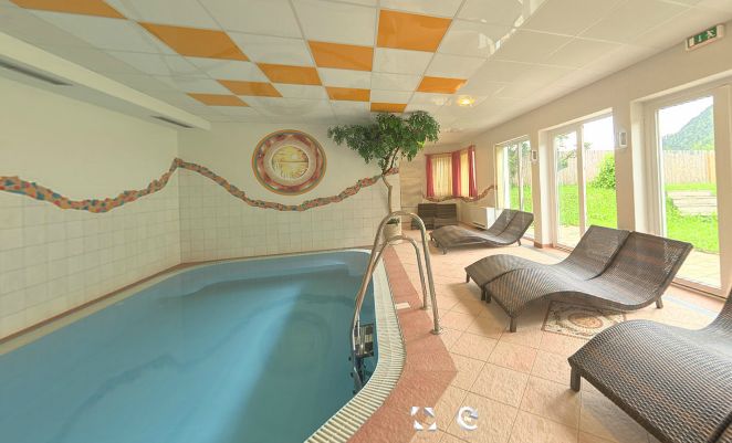 Innenpoolbereich mit kleinem Schwimmbecken, gefliestem Boden, mehreren Korbsesseln, Topfbaum, Wandkunst und großen Fenstern, die viel natürliches Licht hereinlassen.