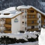 Ein großes, verschneites Alpenhotel aus Holz mit Balkonen und schrägem Dach, umgeben von schneebedeckten Bäumen und Bergen im Hintergrund.