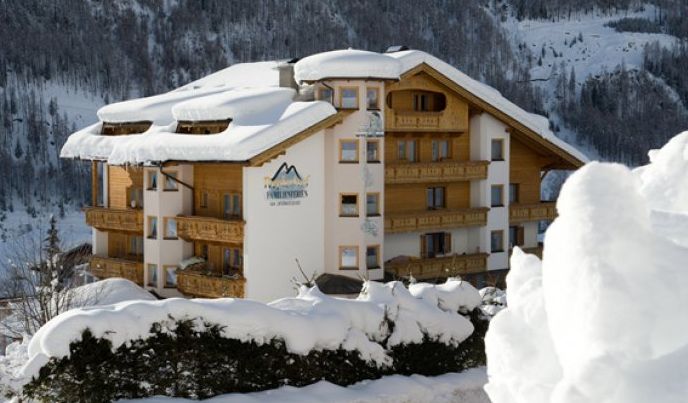 Ein großes, verschneites Alpenhotel aus Holz mit Balkonen und schrägem Dach, umgeben von schneebedeckten Bäumen und Bergen im Hintergrund.