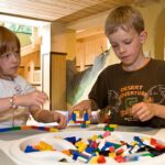 Zwei Kinder sitzen an einem Tisch und bauen mit bunten Plastikklötzen in einem Spielzimmer mit Holzmöbeln und einer gemütlichen Atmosphäre.