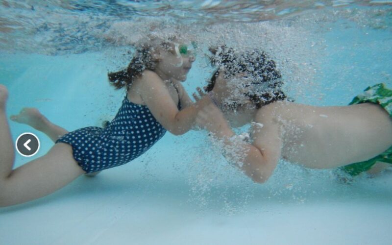 Zwei Kinder in Badeanzügen und mit Schwimmbrillen schwimmen in einem Becken unter Wasser, einander zugewandt und mit Luftblasen um ihre Gesichter.