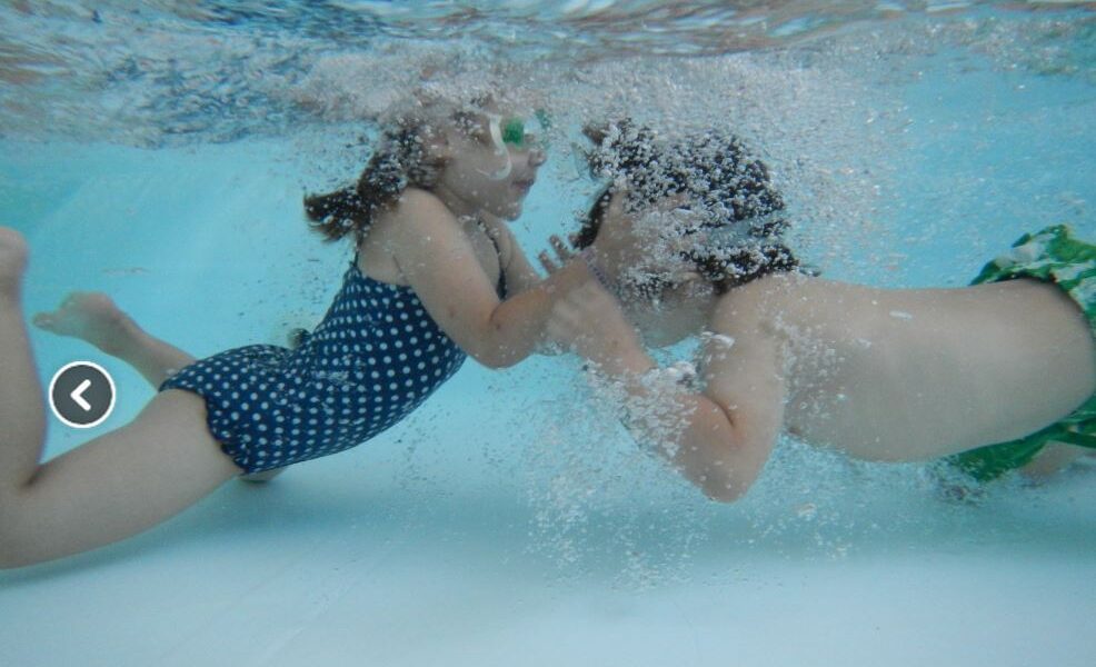 Zwei Kinder in Badeanzügen und mit Schwimmbrillen schwimmen in einem Becken unter Wasser, einander zugewandt und mit Luftblasen um ihre Gesichter.