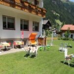 Ein grasbewachsener Hof mit Liegestühlen, einer Rutsche, Tischen mit roten Sonnenschirmen, einem kleinen Spielhaus und einem Gebäude mit Balkonen; im Hintergrund sind die Berge zu sehen.