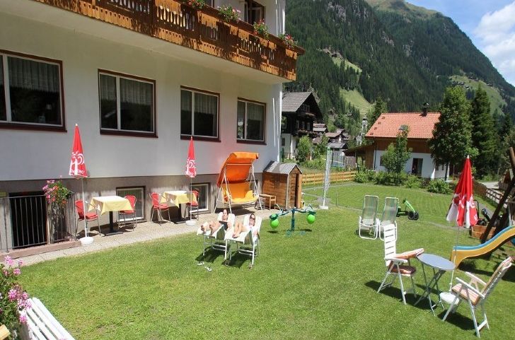 Ein grasbewachsener Hof mit Liegestühlen, einer Rutsche, Tischen mit roten Sonnenschirmen, einem kleinen Spielhaus und einem Gebäude mit Balkonen; im Hintergrund sind die Berge zu sehen.