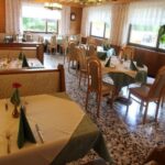 Ein Restaurant mit Tischen, die mit cremefarbenen und grünen Tischtüchern bedeckt sind und jeweils mit Geschirr, Servietten und einer einzelnen roten Blume in einer Vase ausgestattet sind. Große Fenster lassen viel natürliches Licht herein.