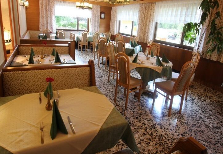 Ein Restaurant mit Tischen, die mit cremefarbenen und grünen Tischtüchern bedeckt sind und jeweils mit Geschirr, Servietten und einer einzelnen roten Blume in einer Vase ausgestattet sind. Große Fenster lassen viel natürliches Licht herein.