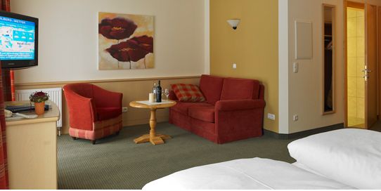 Hotelzimmer mit einem Doppelbett, einem roten Sessel und einem Sofa, einem kleinen runden Tisch mit Flaschen, einem Fernseher auf einer Kommode, Wandkunst und einer offenen Tür, die zu einem Badezimmer führt.