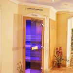 Ein Wellness-Spa-Bereich mit Glastüren, die zu einem Infrarotraum und einer Sauna führen, Zimmerpflanzen, Wandhaken und dekorative Elemente in einem sanft beleuchteten, gefliesten Flur.