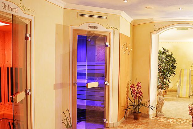 Ein Wellness-Spa-Bereich mit Glastüren, die zu einem Infrarotraum und einer Sauna führen, Zimmerpflanzen, Wandhaken und dekorative Elemente in einem sanft beleuchteten, gefliesten Flur.