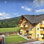 Vierstöckiges gelbes Gebäude mit Balkonen und Blumenkästen, angrenzend an eine grüne Wiese und einen Spielplatz, vor einer Kulisse aus Hügeln und blauem Himmel.