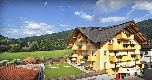 Vierstöckiges gelbes Gebäude mit Balkonen und Blumenkästen, angrenzend an eine grüne Wiese und einen Spielplatz, vor einer Kulisse aus Hügeln und blauem Himmel.