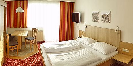 Ein Hotelzimmer mit einem Doppelbett, einem hölzernen Kopfteil, einem an der Wand montierten Fernseher, einem kleinen Tisch mit zwei Stühlen, einem roten Teppich und gestreiften Vorhängen vor einem großen Fenster.