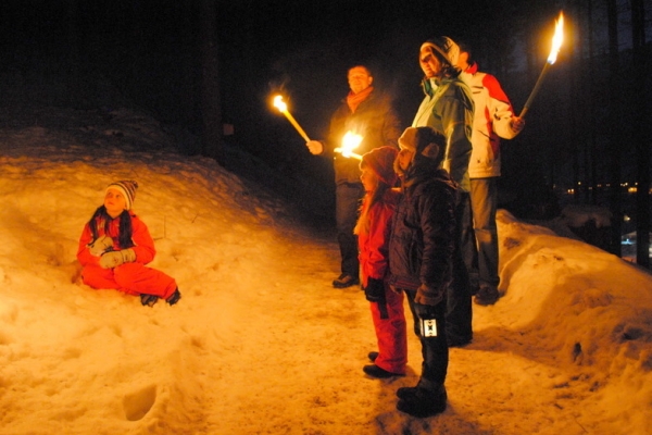 Eine Gruppe von Menschen steht mit Fackeln in der Hand nachts auf verschneitem Boden, während eine Person in der Nähe im Schnee sitzt.