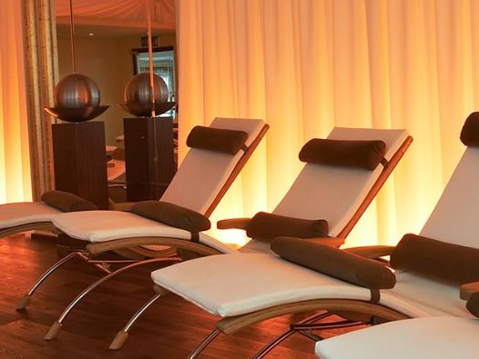 Drei moderne Loungesessel mit weißen Polstern und braunen Kissen sind vor einem sanft beleuchteten, beigen Vorhang aufgereiht. Zwei dekorative Kugeln stehen auf Ständern im Hintergrund.