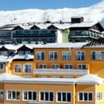 Eine Gruppe farbenfroher Gebäude mit schneebedeckten Dächern steht vor der Kulisse verschneiter Berge unter einem klaren Himmel.