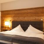 Ein Hotelzimmer mit einem Doppelbett, einem grau gepolsterten Kopfteil, einer Akzentwand aus Holz, zwei Nachttischen und zwei beleuchteten Lampen.