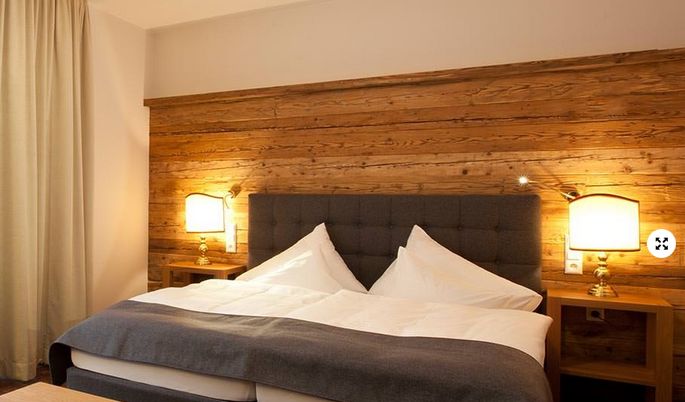 Ein Hotelzimmer mit einem Doppelbett, einem grau gepolsterten Kopfteil, einer Akzentwand aus Holz, zwei Nachttischen und zwei beleuchteten Lampen.