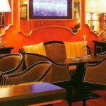 Eine gemütliche Sitzecke mit gepolsterten Stühlen, kleinen Tischen, beleuchteten Lampen und einem Fernseher an einer rot-orangen Wand.