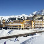 Ein großer gelber Hotelkomplex liegt auf einem verschneiten Hügel mit Bergen im Hintergrund; Menschen gehen in der Nähe des Gebäudes spazieren und Autos sind entlang der Straße geparkt.