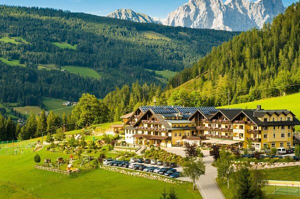 Ein großes Alpenhotel mit Sonnenkollektoren liegt in einem grünen Tal, umgeben von bewaldeten Hügeln und fernen Bergen, unter einem klaren blauen Himmel.