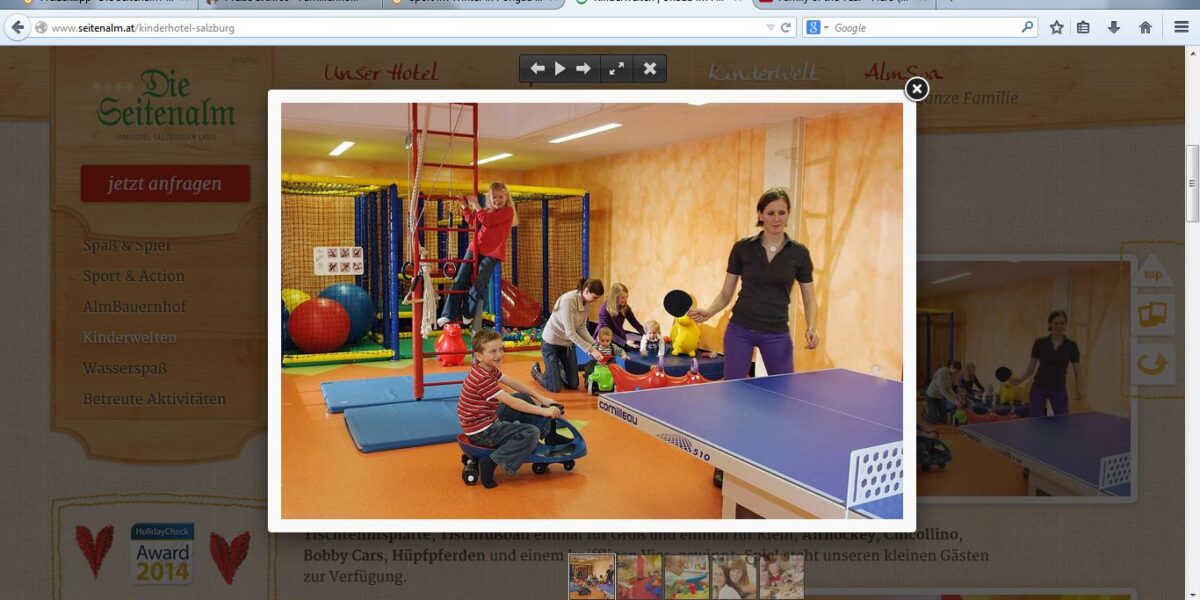 Kinder spielen auf Matten und fahren mit Spielzeug in einem Indoor-Spielbereich, während eine Frau neben einer blauen Tischtennisplatte im Vordergrund steht.