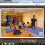 Kinder spielen auf Matten und fahren mit Spielzeug in einem Indoor-Spielbereich, während eine Frau neben einer blauen Tischtennisplatte im Vordergrund steht.